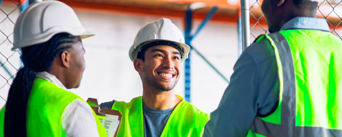 Three workers in safety vests and hard hats smiling in a warehouse Three workers in safety vests and hard hats smiling in a warehouse